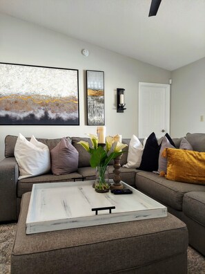 A living room with a grey sofa, a white coffee table, and a vase with flowers. There are two framed artworks on the wall and a white door.