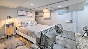 A modern bedroom with a large bed, a grey leather sofa, a bedside table with a lamp, and a wall-mounted shelf.