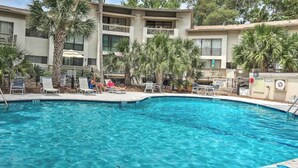 Une piscine entourée de chaises longues et de palmiers, le tout bordé d’un bâtiment doté de balcons et d’une entrée sécurisée par un portail.