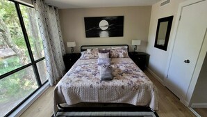 Une chambre à coucher comprenant un lit, deux tables de chevet, une commode, une fenêtre avec des rideaux, un tableau accroché au mur et une porte.