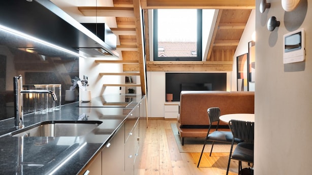A modern kitchen with a black countertop, stainless steel sink, and wooden beams. There is a dining area with a brown sofa and black chairs. - Elegant Downtown Apartment by Vacationy (Porto)