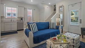 A living room with a blue sofa, a coffee table with a nautical theme, and a kitchen area with white cabinets and a staircase. - Get Happy (Kissimmee)