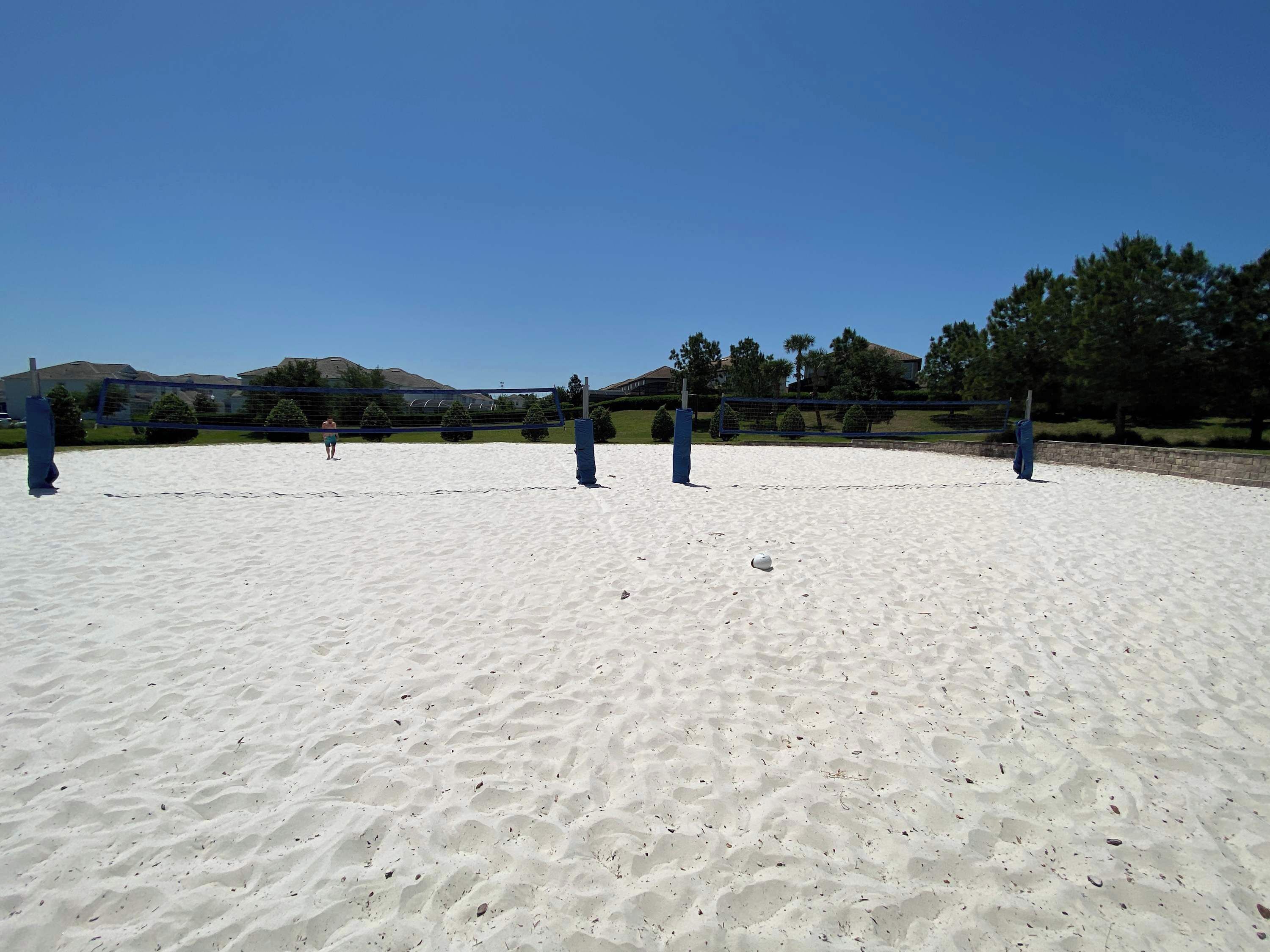 Ein sandiger Strand mit einem Volleyballnetz, Bäumen und Häusern im Hintergrund.