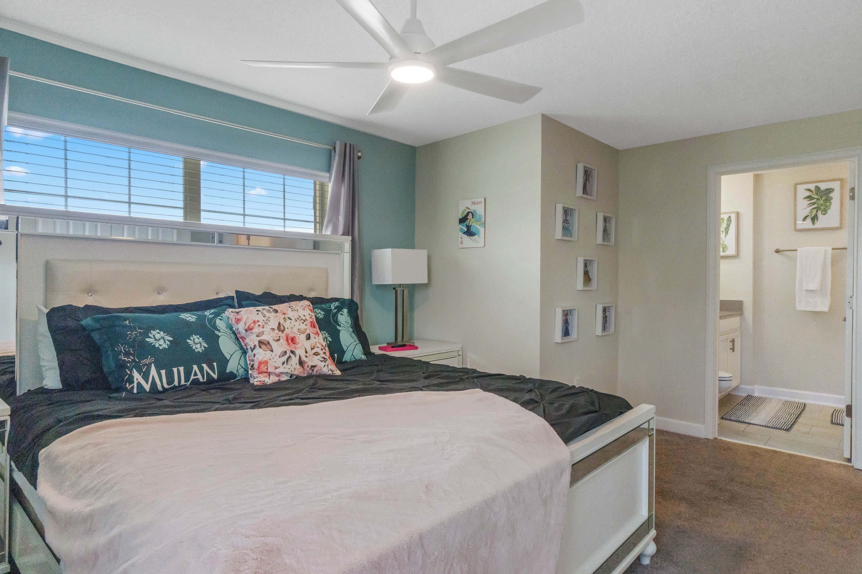 A bedroom with a bed, a ceiling fan, a window with blinds, and a door leading to a bathroom.