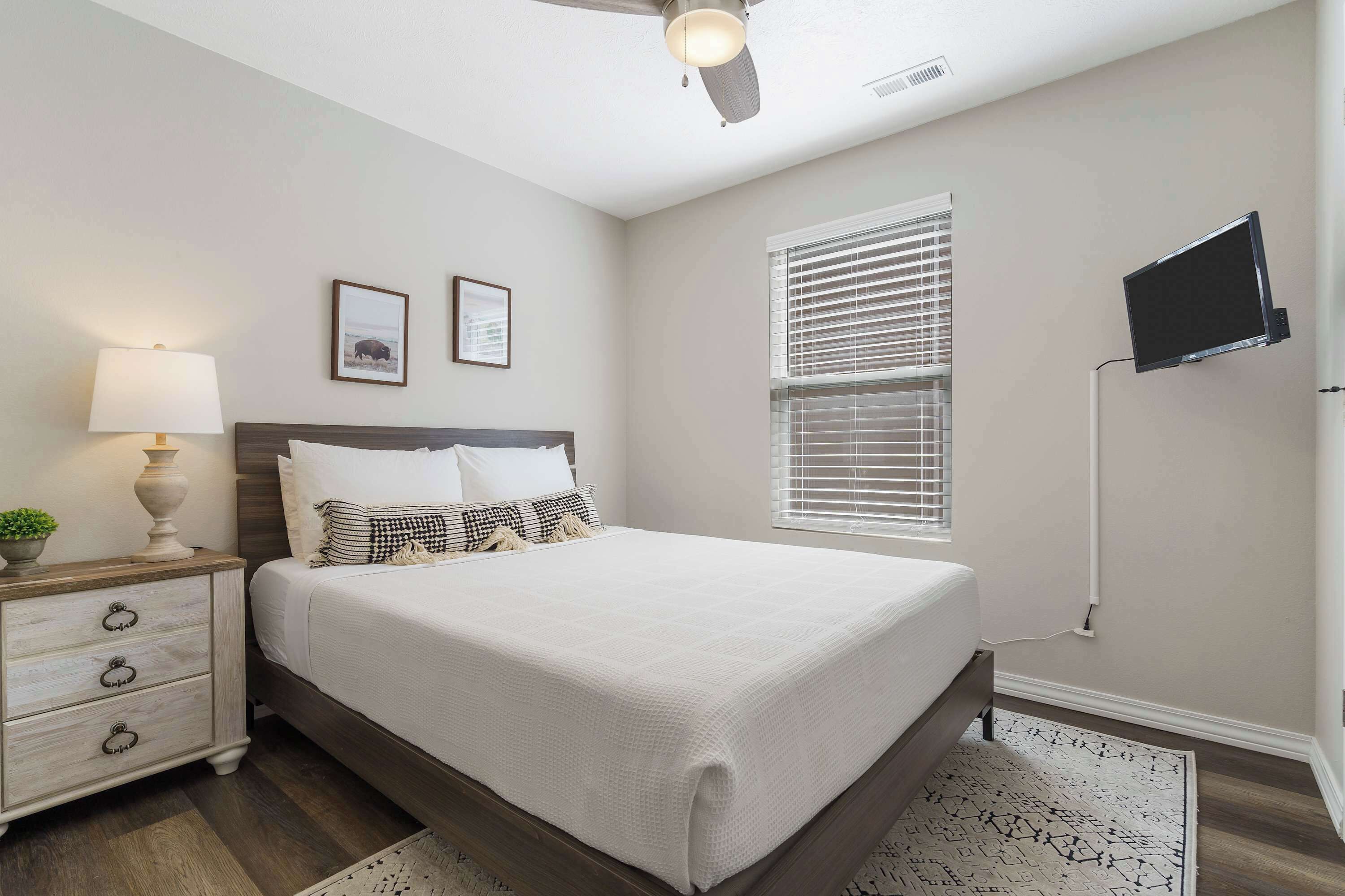 Un dormitorio con cama, mesita de noche, lámpara, televisor montado en la pared y ventana con persianas.