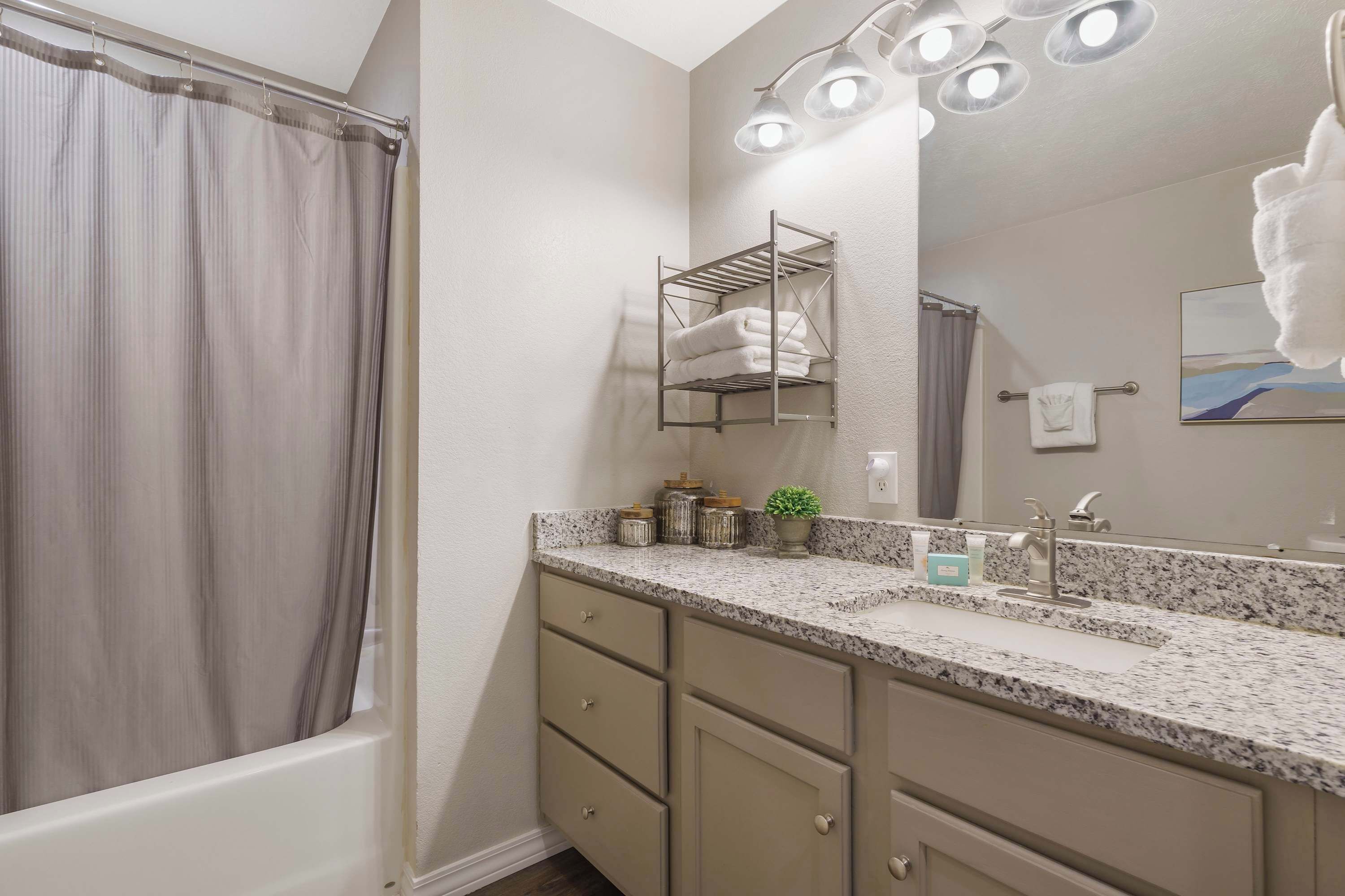Une salle de bain avec un rideau de douche, un porte-serviettes, un lavabo avec un miroir et un plan de toilette avec des pots et une plante.