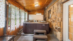 Een hottub in een houten blokhut met stenen muren, een houten plafond en een raam met bloemen gordijnen.
