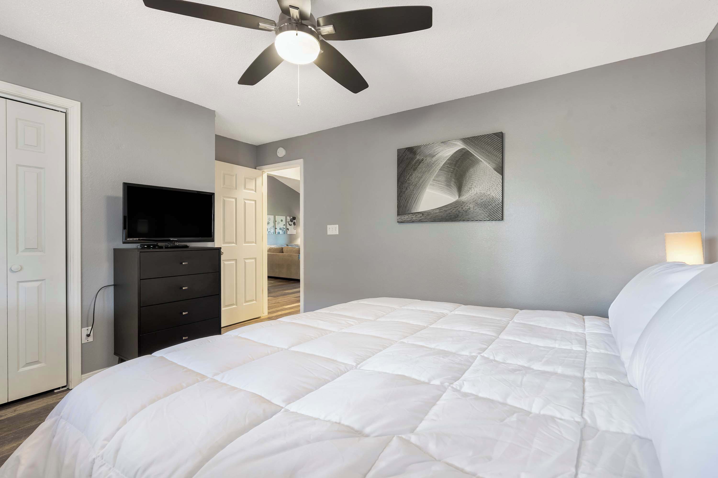 A bedroom with a bed, a ceiling fan, a TV on a dresser, and a door leading to another room.