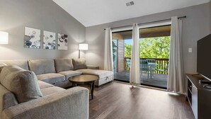 Een moderne woonkamer met een hoekbank, een klein rond tafeltje en een flatscreen televisie. Er is een glazen schuifdeur die leidt naar een terras buiten met een tafel en stoelen.