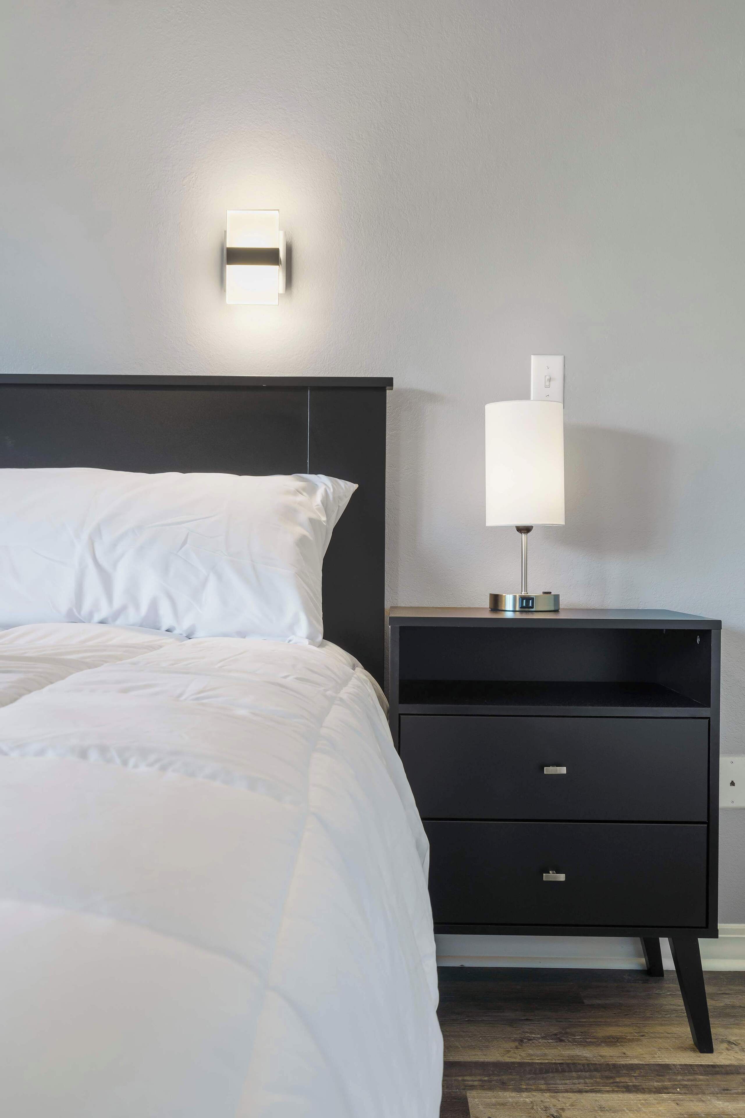 Ein ordentlich bezogenes Bett mit weißer Bettwäsche, ein schwarzer Nachttisch mit Lampe und eine Wandleuchte.