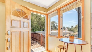 A room with a wooden door, a small table with a stool, and a large window with a view of the outside. - Quaint Carmel - Bungalow Close to Beach (Carmel)