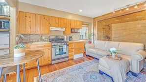 A modern kitchen with wooden cabinets, a built-in oven, and a microwave. There is a dining area with a round table and chairs, a sofa, and a decorative rug. - Quaint Carmel - Bungalow Close to Beach (Carmel)