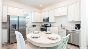 A modern kitchen with white cabinets, stainless steel appliances, and a round dining table set for four. - Shake Shack Heated Pool & Hot Tub (Kissimmee)