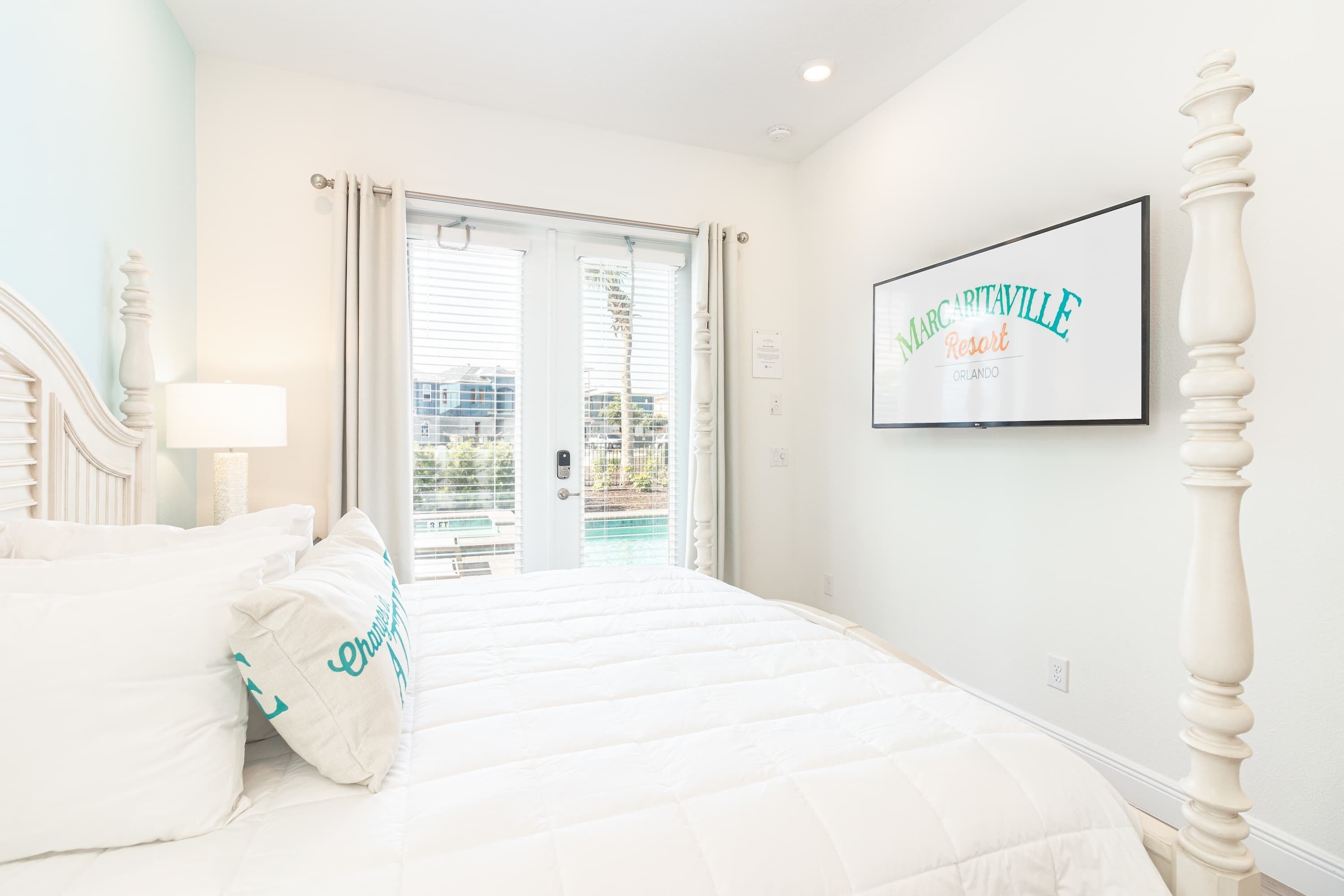 A hotel room with a bed, a nightstand, a lamp, a window with blinds, and a framed sign on the wall.