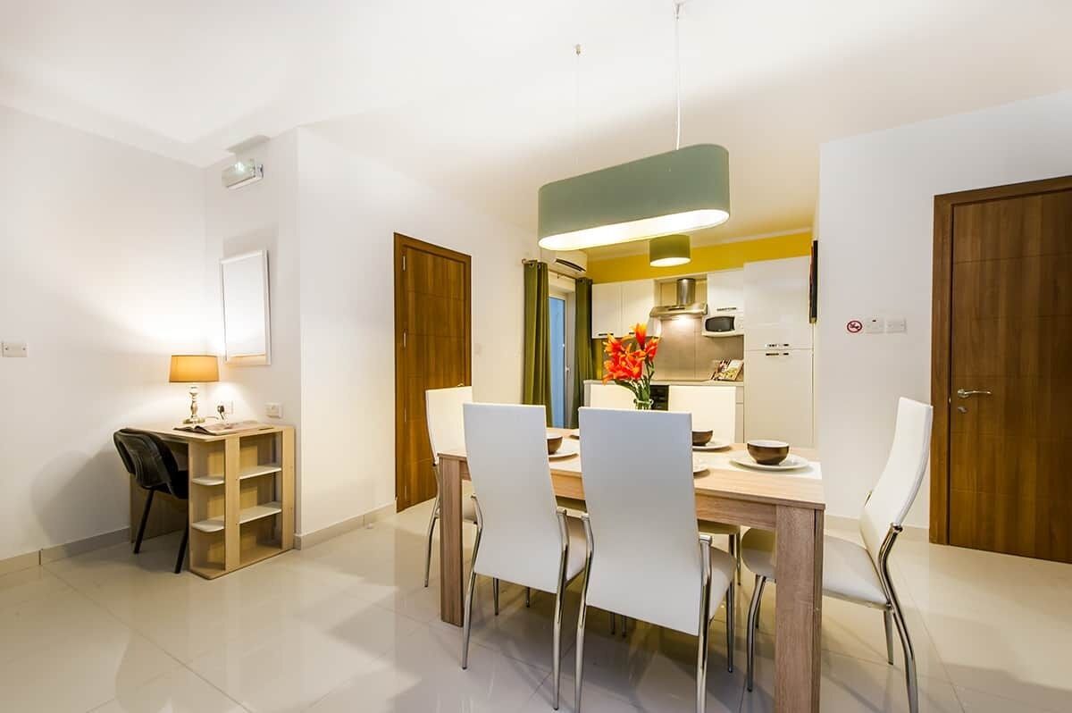 Un comedor moderno con una mesa de madera, sillas blancas, una lámpara colgante y una cocina al fondo.