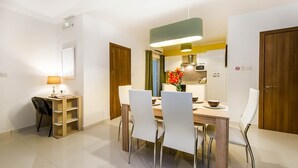Un espace repas moderne avec une table en bois, des chaises blanches, un luminaire suspendu et une cuisine en arrière-plan.