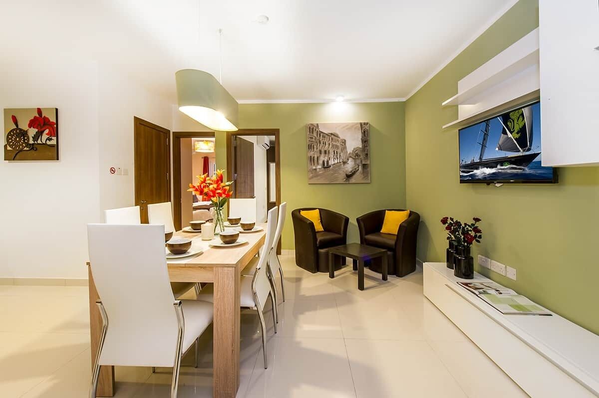 Uma área de refeições com mesa de madeira e cadeiras brancas, uma pequena área de estar com dois sofás e uma televisão fixada na parede.