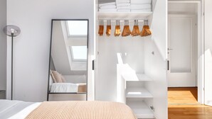 A modern bedroom with a skylight, a bed with a patterned bedspread, a wardrobe with hanging clothes, and a white bathroom with a shower.