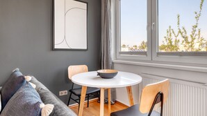 A modern living room with a round white table, two chairs, a grey sofa, and a large window with a view of trees and buildings.