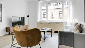 Een moderne woonkamer met een bank, een stoel, een televisie, een tafel en een raam met uitzicht op gebouwen.
