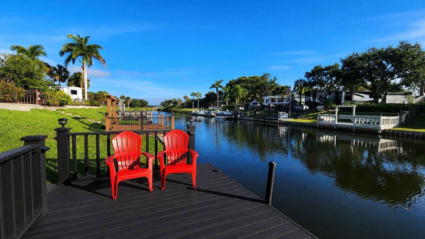 A waterfront deck with two red chairs, a calm river, and houses in the background.