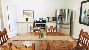 A dining area with a wooden table, chairs, a kitchen in the background, and a refrigerator.