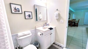 Ein Badezimmer mit Toilette, Waschbecken und Spiegel. An den Wänden befinden sich Bilder in Rahmen und ein Handtuch hängt an der Tür.