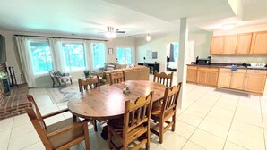 A spacious living room with a large wooden dining table surrounded by chairs, a kitchen area with wooden cabinets, and a view of the outdoors through large windows.