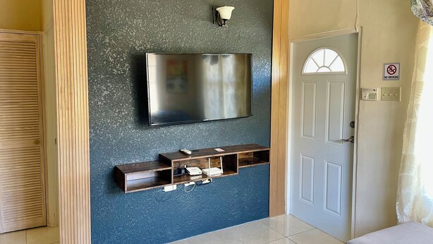 A room with a mounted TV, a wooden shelf with electronic devices, a door with a 'No Smoking' sign, and a patterned curtain.