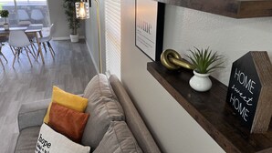 Un salon moderne avec un canapé gris, une table basse et une bibliothèque. On aperçoit une plante en pot sur la bibliothèque et un tableau encadré au mur.