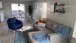 Un salon moderne avec un canapé gris, un fauteuil bleu et une petite table basse. On aperçoit à l’arrière-plan un coin repas avec une table et des chaises.