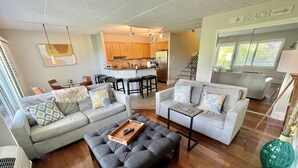 Uma sala de estar moderna com um sofá cinzento, uma poltrona cinzenta e um banquinho a combinar. Há uma mesa de centro de madeira e uma pequena mesinha de apoio. O chão é de madeira e há uma janela grande com persianas. Uma área de cozinha é visível ao fundo, com armários de madeira e uma geladeira.