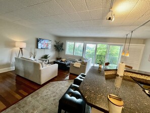 Un salón espacioso con un ventanal grande, un televisor de pantalla plana, una zona de comedor con una barra alargada y una cocina con salpicadero de azulejos.