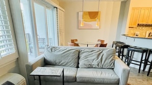 Uma sala de estar moderna com um sofá cinzento, uma mesa de centro de madeira com xícaras, e uma área de refeições ao fundo.