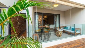 A modern outdoor seating area with a glass balustrade, wooden deck, and a view into a dining space with tables and chairs.