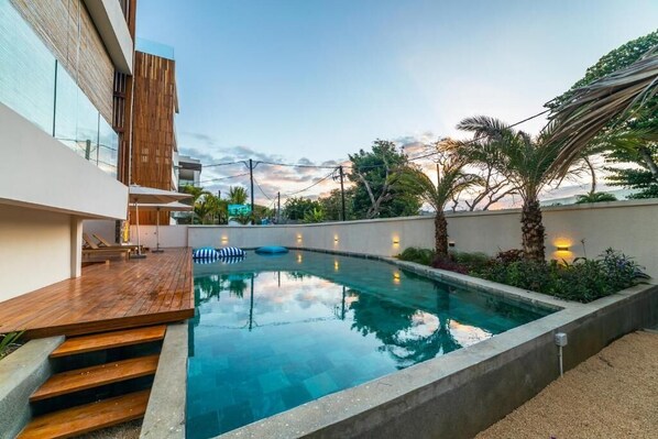A modern pool area with a wooden deck, a concrete pool, and a view of a building with large windows. - Immaculate 2-Bed Apartment in Pereybere Grand Baie (Grand Baie)