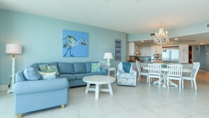 A living room with a sofa, coffee table, and chairs, a dining area with a table and chairs, and a kitchen in the background. - Three Bedroom Condo with Gorgeous Beach Views! Complimentary Boat Slip! (Orange Beach)