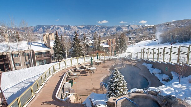 Un espacio exterior cubierto de nieve con asientos y un jacuzzi, rodeado de edificios y una montaña al fondo.