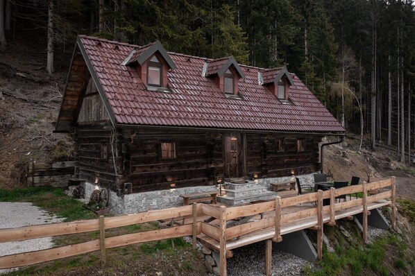 Eine rustikale Holzhütte mit steilem Dach, mehreren Fenstern und einer Holzterrasse mit Sitzgelegenheiten.