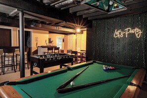Ein Billardtisch mit einem Neon-Schild, das 'Let's Party' beschreibt, in einem Raum mit einem Tischfußballspiel und Barhocker.