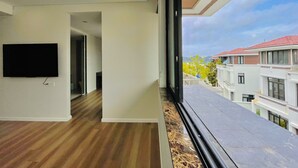 A modern interior with a flat-screen TV mounted on the wall, a wooden floor, and a large window offering a view of neighboring buildings and trees.