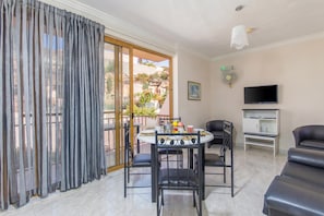A living area with a dining table, chairs, a sofa, a TV, and a balcony with curtains.