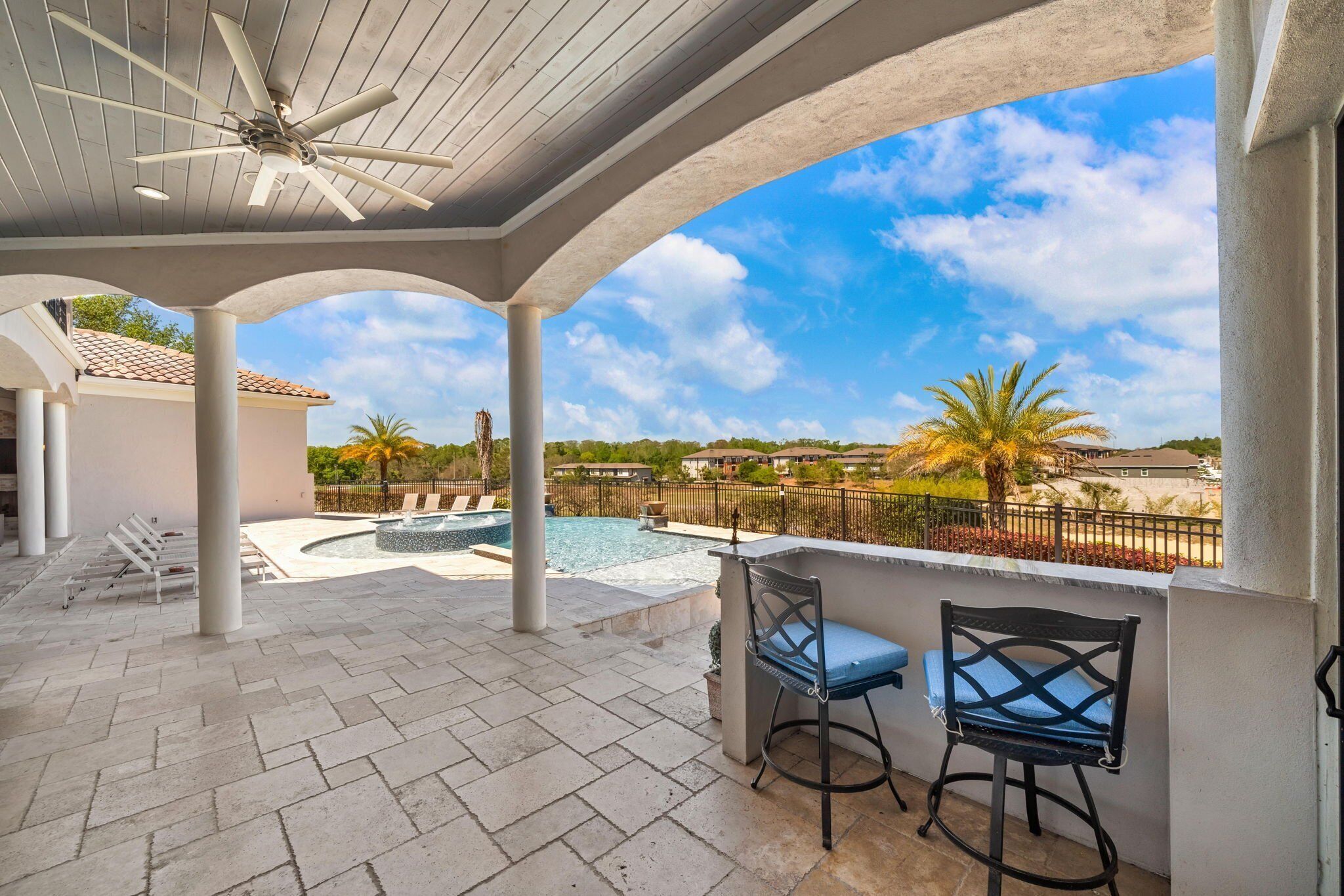 A covered outdoor area with a pool, two bar stools, and a ceiling fan.