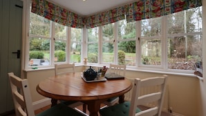 Una mesa redonda de madera con sillas, una ventana con cortina de flores, y vista a follaje exterior.