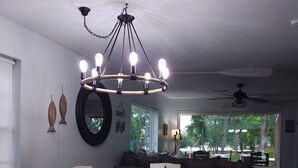 Ein Essbereich mit einem dunklen Holztisch und weißen Stühlen, einer Kronleuchter mit mehreren Lampen sowie ein Blick ins Freie durch ein großes Fenster.