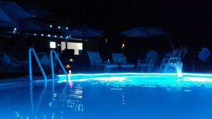 Ein Nachtbild eines Schwimmbeckens mit beleuchteten Rändern und einer Wasserspielanlage.