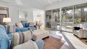 Een ruime woonkamer met een grote schuifpui naar een terras, een blauwe zithoek, een beige poef en een gestreepte fauteuil.