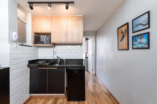 A modern kitchen with black cabinets, a black countertop, and a built-in microwave. There are wooden cabinets above the microwave and a refrigerator to the right. The kitchen has white subway tiles and a wooden floor.