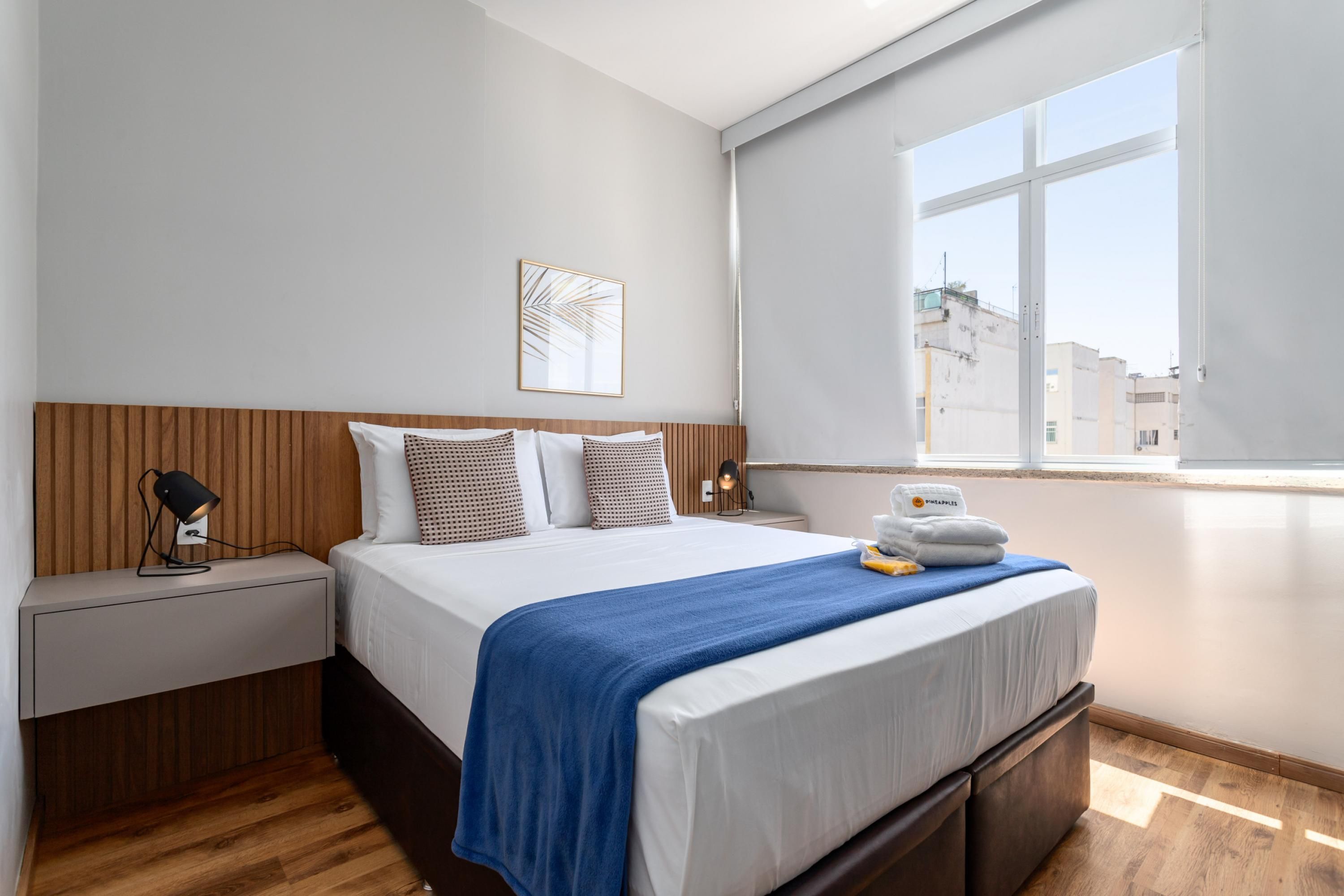 A modern hotel room with a large bed, a bedside table with a lamp, a window with a view of buildings, and a wall-mounted artwork.