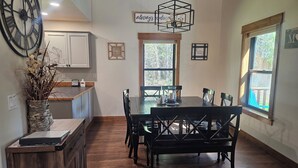 A dining area with a wooden table and chairs, a large clock, and a view of the outdoors through a window.